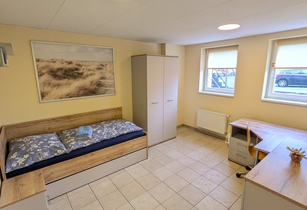 Auf dem Bild ist ein Zimmer mit einem Bett, einem Nachttisch, einem Kleiderschrank, einer Kommode, einem Schreibtisch, einem Stuhl sowie einem Bild an der Wand zu sehen.