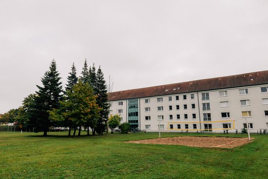 Eine Wiese mit einem Volleyballfeld und Bäumen, im Hintergund das viergeschossige Wohnheim in der Geschwister-Scholl-Straße.