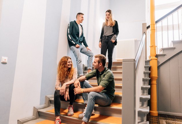 Vier Studierende sitzen und stehen auf der Treppe des Wohnheims im Hausflur, der mit freundlichen, hellen farben gestaltet ist. 