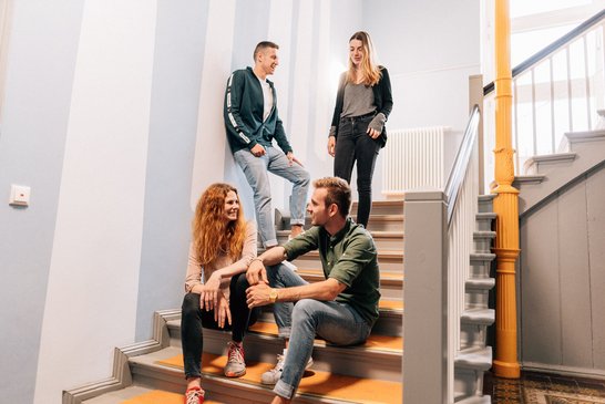 Vier Studierende sitzen und stehen auf der Treppe des Wohnheims im Hausflur, der mit freundlichen, hellen farben gestaltet ist. 