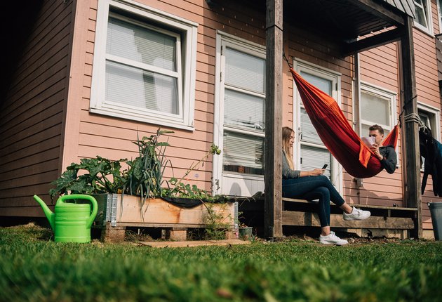 Eine junge Frau sitzt mit einem Handy auf der Terrasse eines orangen Wohnhauses der Wohnheimanlage Holzhausen. Daneben liegt ein junger Mann in einer roten Hängematte und liest. Neben der Terrasse ist ein kleines Beet angelegt, vor dem eine Gießkanne steht.