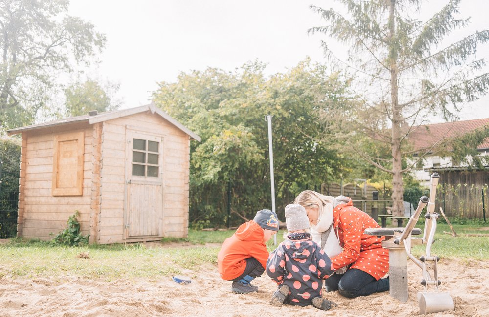 Eine Kita-Erziehrin spielt mit zwei Kindern in einem Sandkasten auf dem Hof des Kindergartens. Hinter dem Sandkasten stehen ein Gartenhäuschen und verschiedene Bäume.