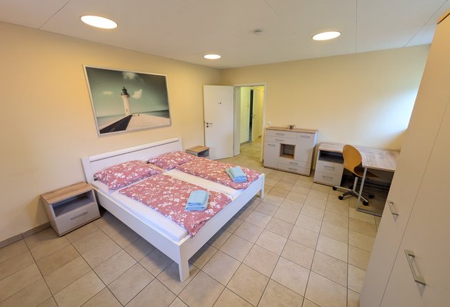 Auf dem Bild ist ein Zimmer mit einem Doppelbett, zwei Nachttischen, einem Kleiderschrank, einer Kommode, einem Schreibtisch, einem Stuhl sowie einem Bild an der Wand zu sehen.