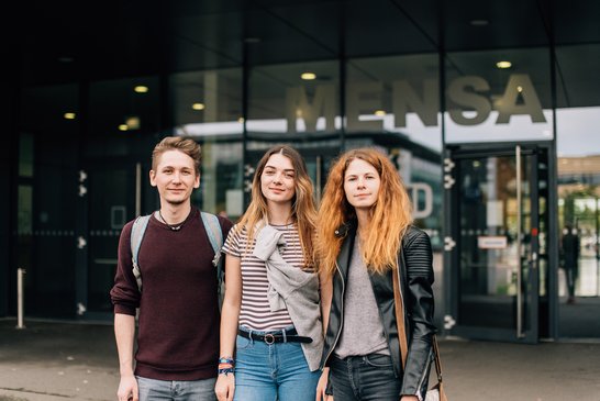 Drei Studierende stehen vor einem Gebäude mit der Aufschrift Mensa