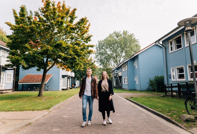 Ein junger Mann und eine junge Frau gehen auf einem Weg zwischen den blauen, zweigeschossigen Häusern der Wohnheimanlage Holzhausen. Vor den Häusern wachsen grüner Rasen und Bäume. An den Hauswänden sind überdachte Fahrradständer.